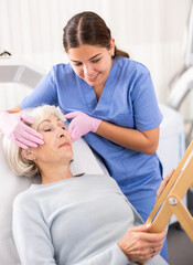 Satisfied senior woman looking at mirror after professional cosmetology treatment with beautician...