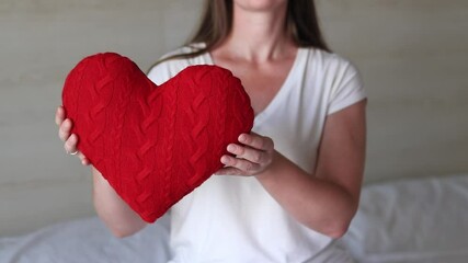 Girl holding knitted red heart, close-up. Valentine's Day. Heart-shaped pillow. Valentine's Day gift. Girl sitting on bed with toy heart. Symbol of love, gift. Romantic young woman with pillow