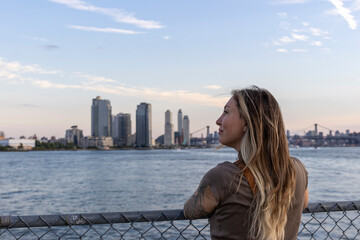 A woman gazes at the New York City skyline during sunset by the river, capturing the vibrant colors and iconic buildings in the evening light