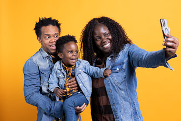 Obraz premium Young family smiling for selfies on smartphone with their boy against yellow background, capturing sweet memories together in studio. Proud african american mom and dad posing with the child.