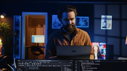 Portrait of smiling man doing IT support job from home, standing in personal office with laptop in...