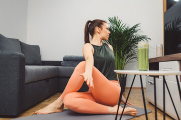 A young woman drinks a vegetable and fruit smoothie in her living room and doing yoga on yoga mat.