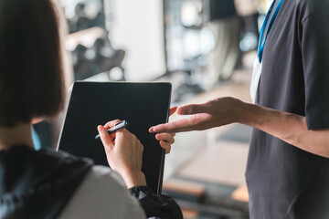 スポーツジムでタブレットにサインする顧客と案内するトレーナーの手元
