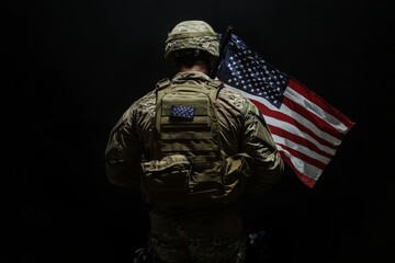 A soldier in camouflage uniform and helmet stands with his back to the camera holding a US flag.