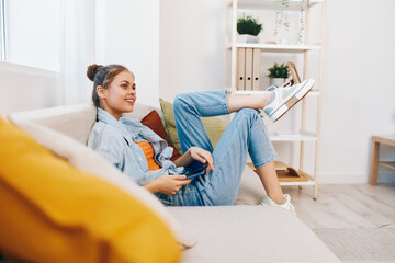Obraz premium Smiling woman holding a mobile phone on a cozy sofa at home, enjoying online games and reading, while taking a cheerful selfie and chatting with friends in cyberspace.