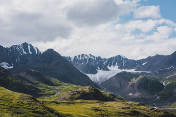 Dramatic scenery in alpine valley with creek among green hills and rocks with view to rocky pointy peak, large snow-capped peaked top, mountain range and big glacier tongue under clouds in blue sky.