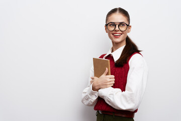 Young girl with glasses smiling while holding a book, wearing a red sweater and white shirt Simple background emphasizes her cheerful expression and academic vibe