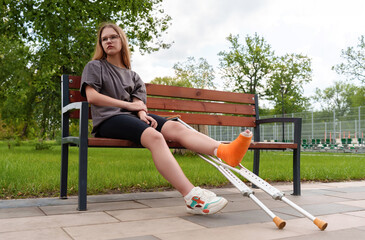 Casual-dressed female adolescent with cast on leg appears disheartened while seated on bench in...