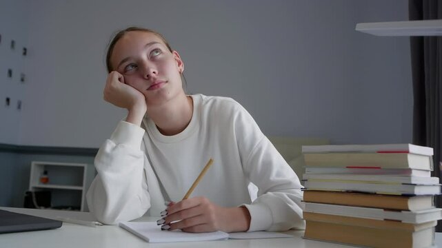 Teenage girl is sitting at her desk with a bored expression, holding a pencil and daydreaming while doing her homework