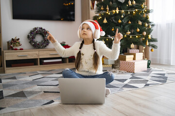 Caucasian girl wearing Santa hat sits on floor listening to music in wireless headphones using laptop near Christmas tree. Celebrates festive season with joy and technology at home.