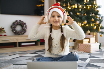 Happy Caucasian girl wearing Santa hat listens to music on wireless headphones while sitting on...