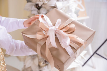 Hands with wrapped christmas gift with white bow on background of christmas tree lights. Stylish present. Merry Christmas and Happy New Year celebration theme.packaging, shopping, delivery concept.