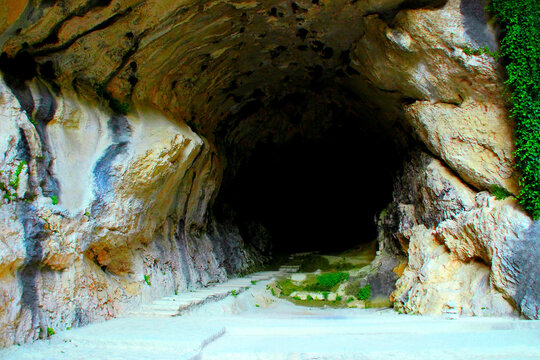 Arcane view with a cave near Eremo di Santa Maria Infra Saxa in Parco Naturale Gola della Rossa e di Frasassi, its engulfing darkness surrounded by furrowed, jaggy, craggy rock, with redeeming green