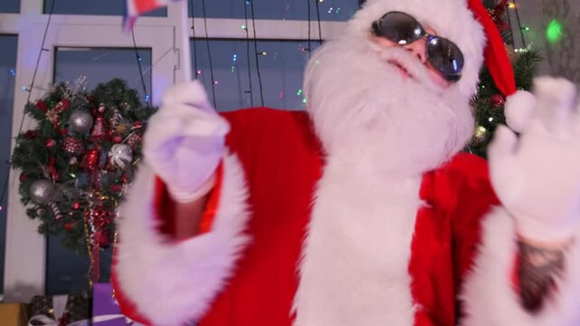 Man in Santa Claus costume dancing and waving an Britain flag, next to Christmas tree and twinkling garlands, during Xmas party.