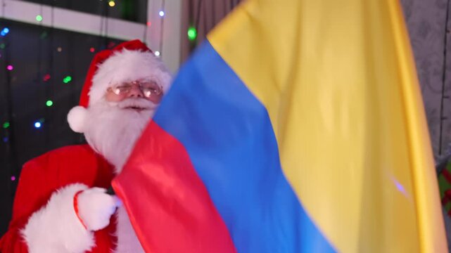 Santa Claus waving Colombian flag holding a heart shaped gift box at Xmas party among festive decorations next to Christmas tree. Colombia support. Happy New Year celebration.