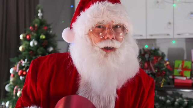 Charismatic whitebearded Santa looking at camera through glasses with gift box heart dancing against background of Christmas tree. Close up.