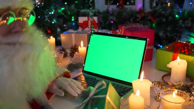 White-bearded Santa in a hat and white gloves sits at a laptop with a chroma key screen surrounded by Christmas decorations, lit candles, a festive tree and gifts.