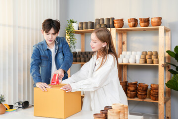 Two young entrepreneurs teamwork of Asian man and Caucasian woman packing a large box for shipping, preparing handcrafted pottery for delivery in their home studio filled with ceramic products - Powered by Adobe