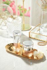 A top-down photo depicts two unlabeled lotion bottles on a brass tray with white flowers, surrounded by glass boxes, jewelry, and gold-colored ornaments, set against a luxurious backdrop of white silk