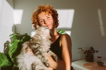portrait red-haired curly young woman with pet gray fluffy cat in arms, lit by bright sun