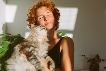 portrait red-haired curly young woman with pet gray fluffy cat in arms, lit by bright sun