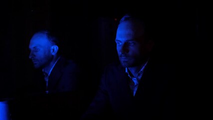 A businessman in a suit sits contemplatively in a dark room. His face is illuminated by a dim blue light reflecting off a mirror, creating a moody and mysterious atmosphere