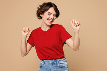 Young smiling happy woman she wear red t-shirt casual clothes doing winner gesture celebrate clenching fists dance isolated on plain pastel light beige background studio portrait. Lifestyle concept.