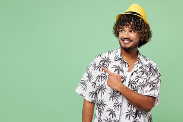 Young happy fun smiling Indian man he wearing beach shirt hat rest near hotel pool point index finger aside on empty area isolated on plain green color background. Summer vacation sea sun tan concept.