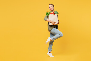 Full body side view young smiling happy woman wear casual clothes hold bag takeaway mock up with food products look aside raise up leg isolated on plain yellow background. Delivery service from shop.