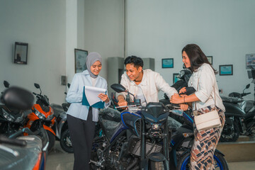 Asian man holding the handlebar of a new motorcycle with his wife and saleswoman looking at the...