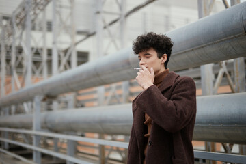 Fashionable young man smoking a cigarette in front of a large industrial building with pipes on the side