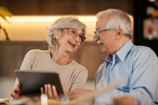 Cheerful senior couple laughing at dining table and holding tablet.