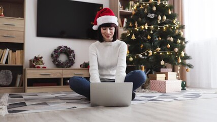 Caucasian woman in twenties wearing Santa hat, sitting with laptop near Christmas tree. Festive home setting includes decorated tree, gifts, and cozy atmosphere.