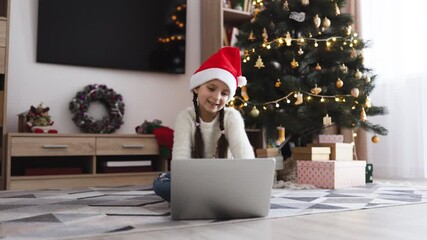 Young Caucasian girl wearing Santa hat sitting on floor engaged in online video call. Christmas tree with decorations and gifts in background creates festive atmosphere. - Powered by Adobe