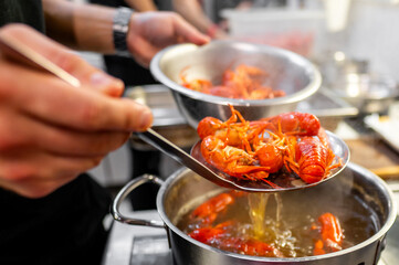 A close-up of hands scooping boiled red crayfish from a steaming stainless steel pot with a slotted spoon. The vibrant crayfish and rising steam suggest a freshly prepared seafood meal