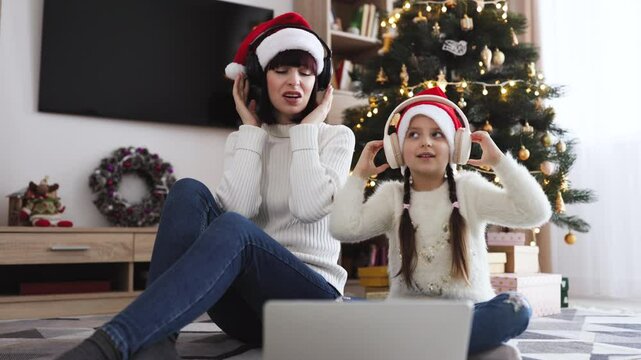 Family leisure during the festive season against the background of Christmas tree. Caucasian young adult mother and little daughter sing songs while listening to carols in headphones using a laptop.