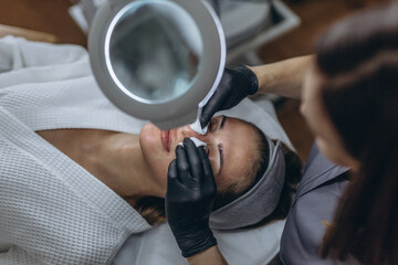 Cosmetologist in rubber gloves performs facial cleansing using a magnifying glass with LED light...