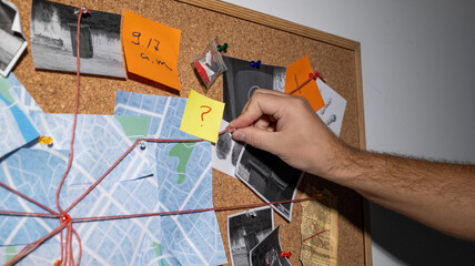 A hand hooks a new clue onto a detective board with a map, photographs and clues marked with red threads connecting at one point.