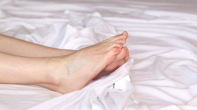 Female legs in bed on white sheets close-up. Female feet. Bare feet of a girl peeking out from under the blanket. Legs wiggling their toes on white bed linen. Rest
