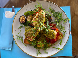 Close-up plate of grilled vegetables and chicken on toast is served in a stylish, bright seaside restaurant. Dish is garnished with fresh arugula and accompanied by a variety of seasonings and