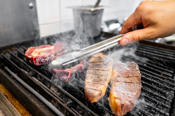 A close-up of a hand using tongs to grill juicy steaks on a barbecue, with smoke rising from the cooking meat. Perfect for summer BBQ, food, and outdoor cooking themes.