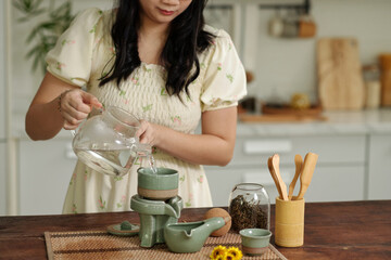 Woman pouring hot water in tea pot while making herbal tea in the kitchen