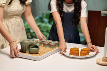 Woman putting tea set on table and her daughter serving baked cakes