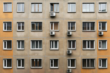 Facade of a residential building featuring multiple rows of windows and air conditioning units attached to several of them, with an alternating color pattern.