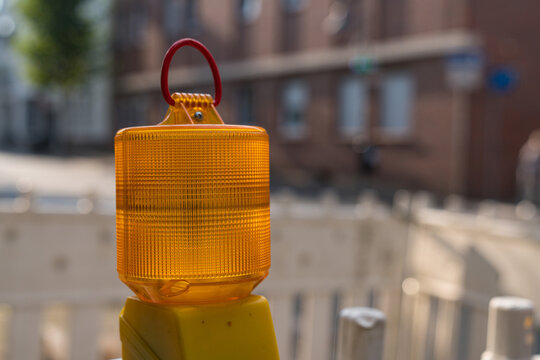 A bright yellow construction light sits atop a post, illuminating a street work area. Surrounding the light, there are signs of ongoing road work and a wooden barrier in the background.