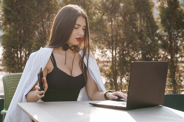 Stylish woman working on laptop outdoors with smartphone in hand surrounded by greenery