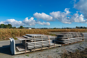 Concrete slabs are neatly stacked on a trailer parked alongside a quiet rural highway. A field of corn stretches out in the background under a clear blue sky.
