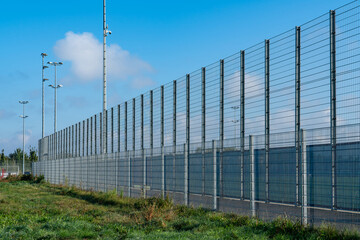 High security fence encompasses a facility, anchored by floodlights, under a bright blue sky. Lush...