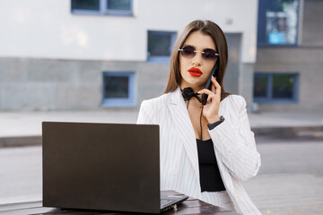 Stylish businesswoman in sunglasses talking on the phone while working on a laptop outdoors