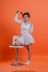 full length young asian woman sexy white sweater holding rope and hair back legs leaning on hydraulic chair, standing on orange background. for fashion, lifestyle, beauty concept